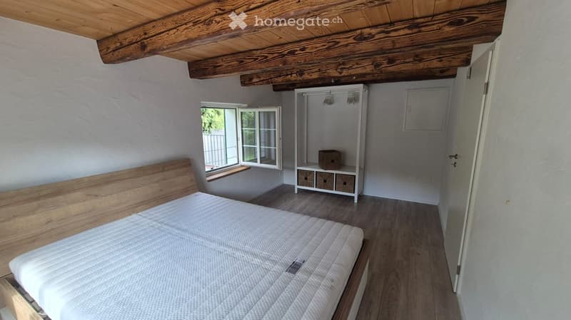 Cozy bedroom with wooden ceiling beams, a large bed with a wooden headboard, a white wardrobe, and a small storage unit. The room has wooden flooring and a window with a view of the garden.
