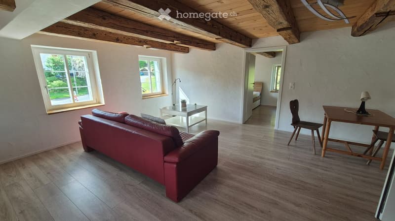 Living room with exposed wooden beams, wooden flooring, red leather sofa, small table, and a chair.