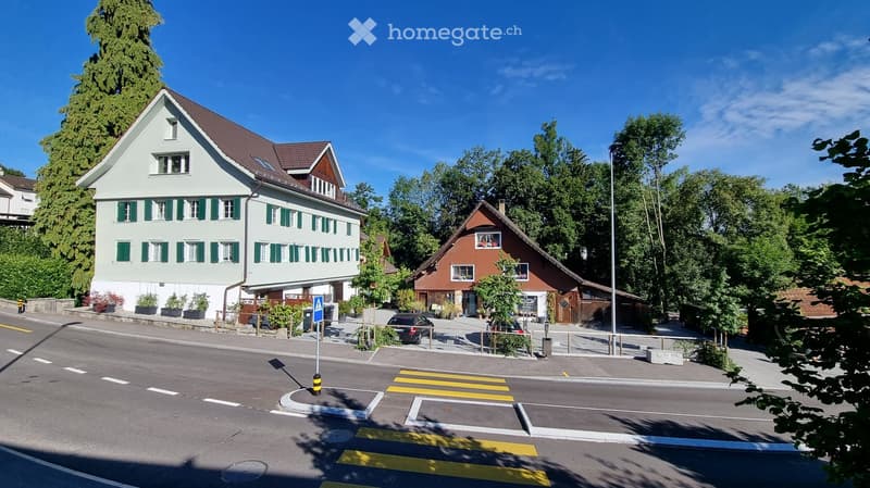 Two-story residential building with green shutters and a brown roof, surrounded by trees and a street crossing.