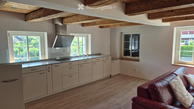 Open-plan kitchen and living area with light-colored cabinets, granite countertop, red leather sofa, and exposed wooden beams on the ceiling.