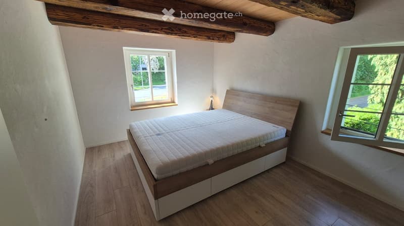 Cozy bedroom with wooden bed, nightstand, and large windows offering a view of greenery.