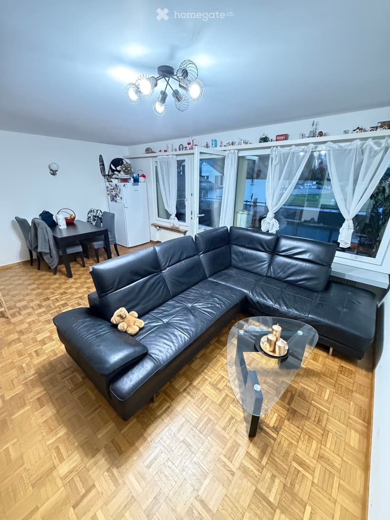 Modern living and dining area with a black leather sectional sofa, glass coffee table, dining table with chairs, and a refrigerator in the background.