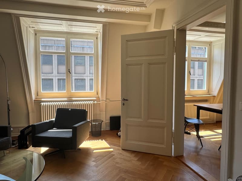 Bright living room with wooden flooring, large windows, a sofa, a chair, a glass coffee table, and a small table in the adjacent room.