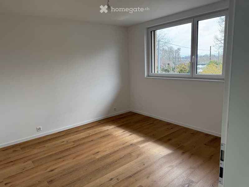 Empty room with wooden flooring, white walls, and large windows offering a view of the outdoors.