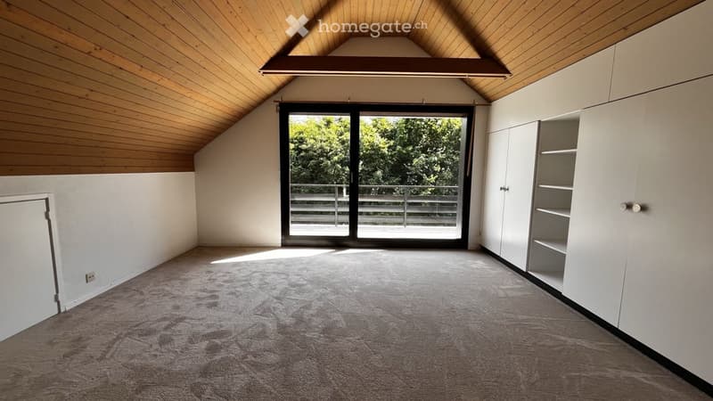 Empty attic room with wooden slanted ceilings, carpeted floor, large sliding glass doors leading to a balcony, and built-in white storage units.