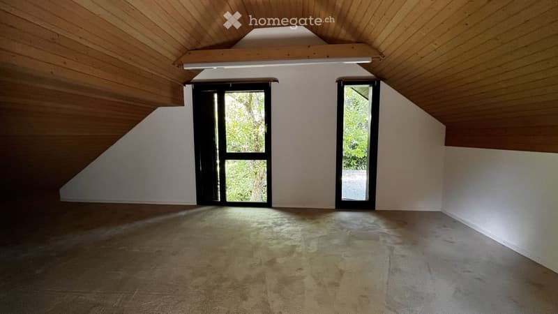 Empty attic room with wooden slanted ceilings, large windows, and concrete floor.