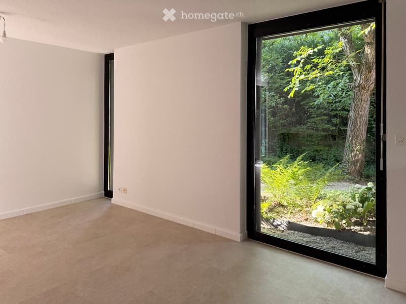Empty room with light-colored walls and wooden flooring, featuring a large black-framed sliding glass door opening to a garden with trees and plants.