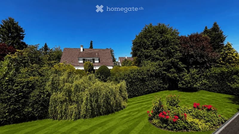 Well-maintained garden with neatly trimmed hedges, a manicured lawn, and vibrant red flowers in a circular bed. A two-story house with a dark roof and white trim is partially visible behind the greenery.