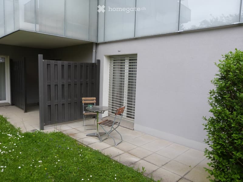 Outdoor patio area with a small table and two chairs, surrounded by a modern building with large windows and a black gate.