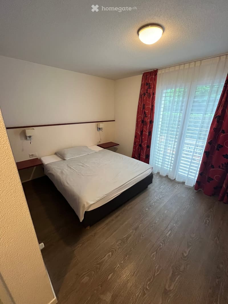 Cozy bedroom with a double bed, nightstands, and red curtains on the window. The room features wooden flooring and a ceiling light.