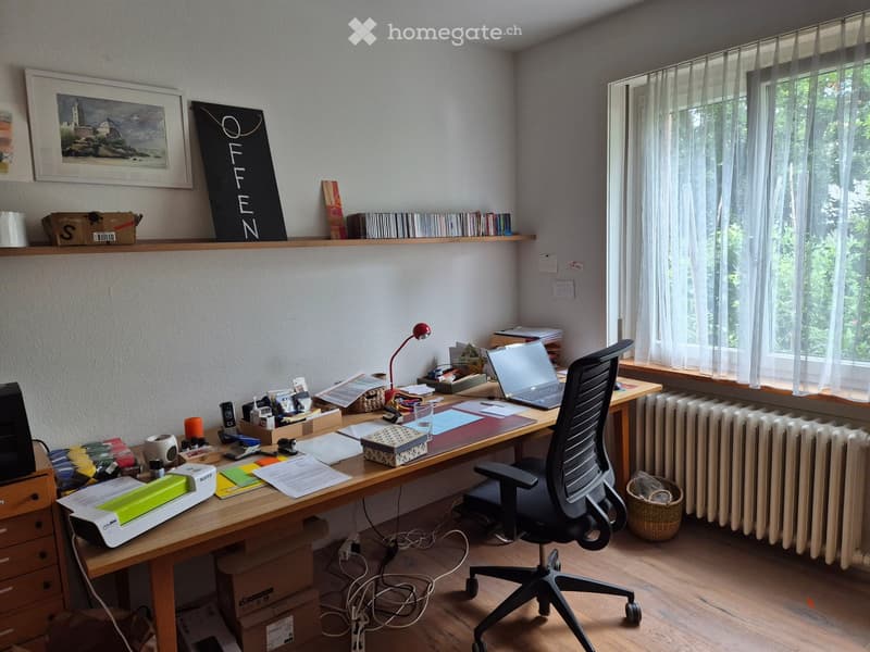 A well-organized home office with a wooden desk, ergonomic chair, laptop, printer, and various office supplies. Shelves hold books, a picture frame, and decorative items. Natural light streams in from a window with sheer curtains.