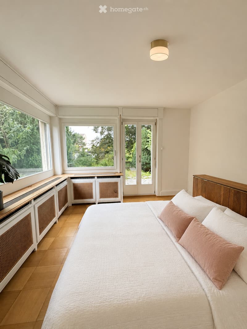 Bright bedroom with wooden flooring, large windows, a neatly made bed with white and pink bedding, a nightstand, radiator, and a plant near the window.