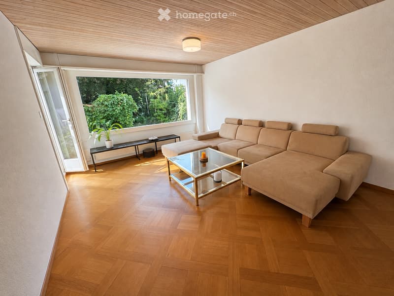 Modern living room with light wooden flooring, a large window with a view of greenery, a beige sectional sofa, a glass coffee table, a side table with a plant, and a small candle on the coffee table.