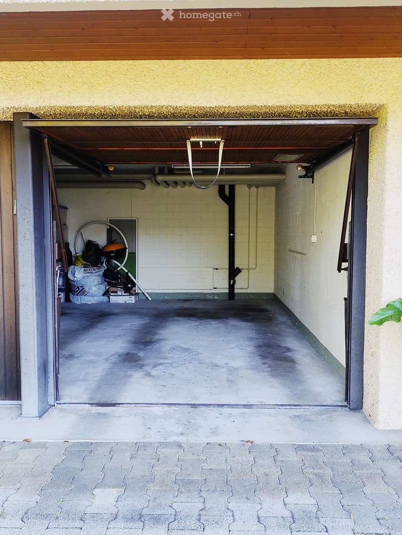 Open garage with concrete floor, white brick walls, and storage items in the corner.