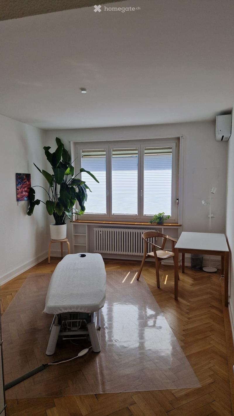 Bright room with wooden flooring, large window with blinds, a massage table, a chair, a small table, a plant on a stool, and a radiator under the window.