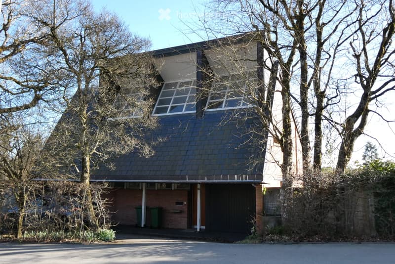 House. Two-story house with a dark roof and large windows, surrounded by trees and greenery.