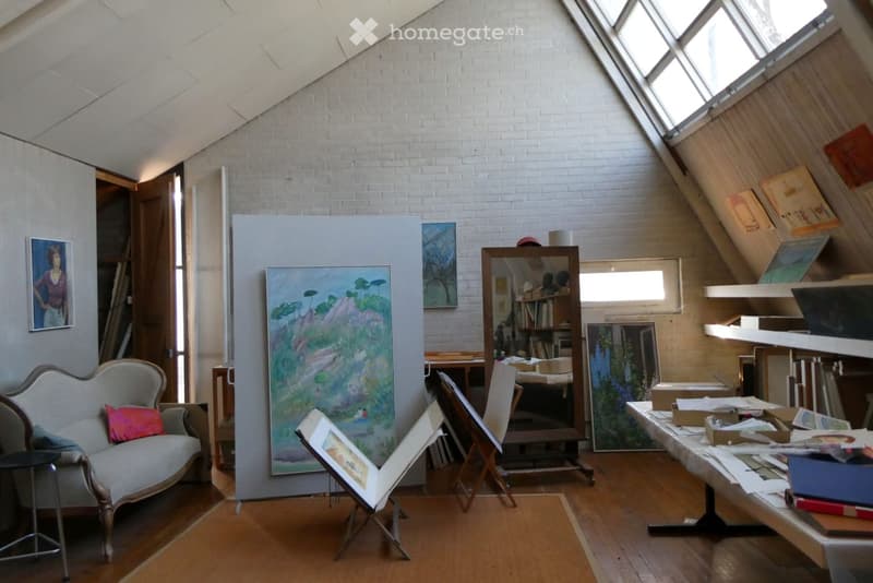 Furnished Room. An attic studio with exposed brick walls, wooden floors, and large skylights. Features a sofa, chair, table, mirror, bookshelf, and several artworks displayed on walls and easels.