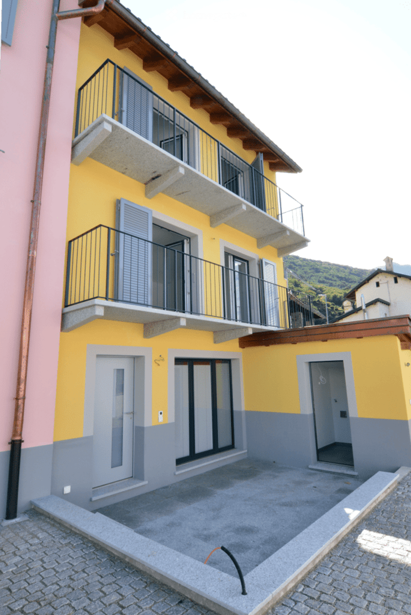 Two-story yellow building with balconies, grey lower facade, and a small paved courtyard.