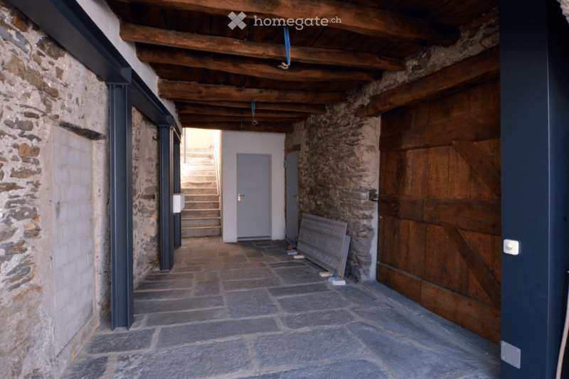 Narrow stone-walled corridor with exposed wooden beams, stone floor, and a staircase leading upwards.