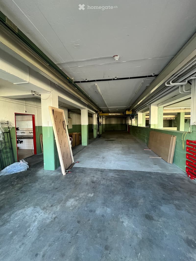 Empty industrial-style corridor with concrete floors, exposed pipes, and minimal furnishings. Green-painted walls, a few scattered items like cardboard boxes and a plastic bag, and a small room visible on the left.