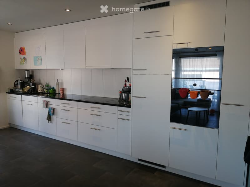 Modern kitchen with white cabinets, black countertops, and stainless steel appliances. Features an oven, coffee maker, blender, and a small stool inside the oven.