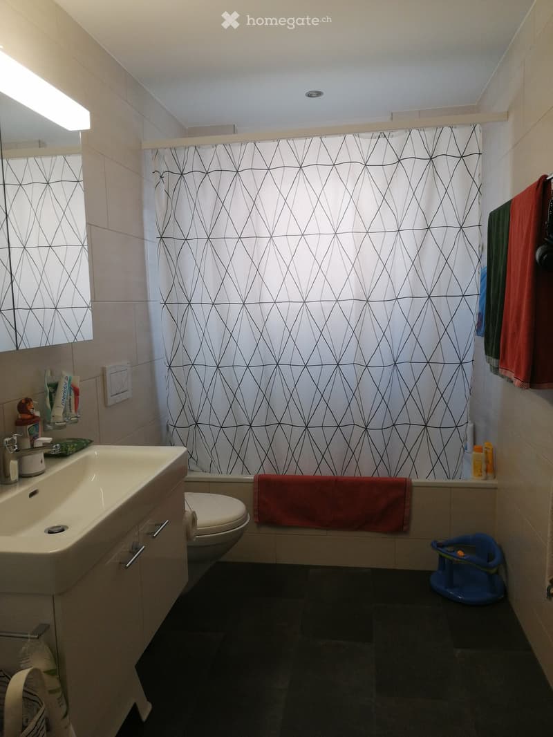 Modern bathroom with a white sink, toilet, mirror, and a geometric-patterned shower curtain. A red towel is draped over the edge of the bathtub, and a blue baby seat is placed on the floor.