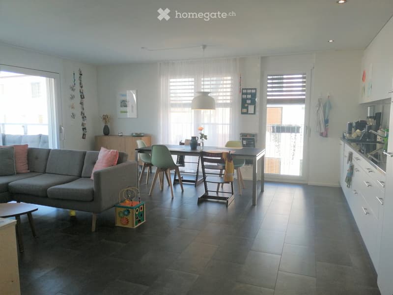 Open-plan living and dining area with a grey sofa, coffee table, dining table with chairs, and a modern kitchen area with cabinets and appliances.