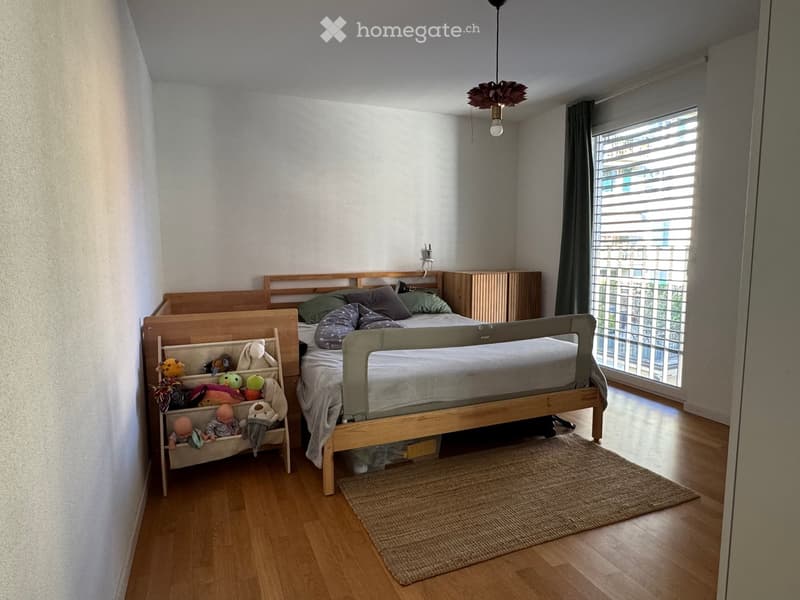 Cozy bedroom with a wooden bed, nightstand, toy basket, and a small rug on the floor. Large window with blinds allows natural light to enter.