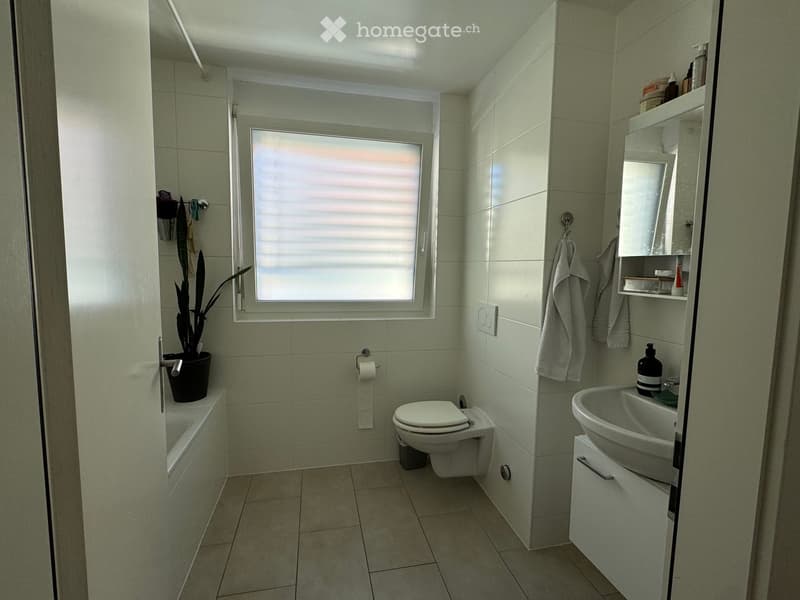 Modern bathroom with tiled walls and floor, featuring a toilet, sink, bathtub, and a small window with blinds. A towel hangs on a rack, and a potted plant sits on the edge of the bathtub.