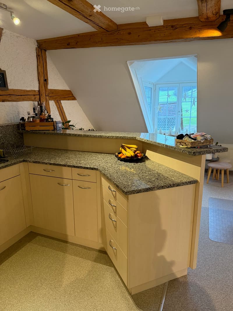 Bright kitchen with light-colored cabinets, granite countertops, and a small window overlooking the garden. The counter has a bowl of fruit, a basket, and a few bottles.