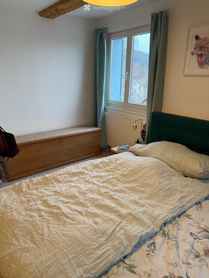 Cozy bedroom with a large bed, nightstand, lamp, and a window with curtains. A wooden chest and a framed picture are also visible.