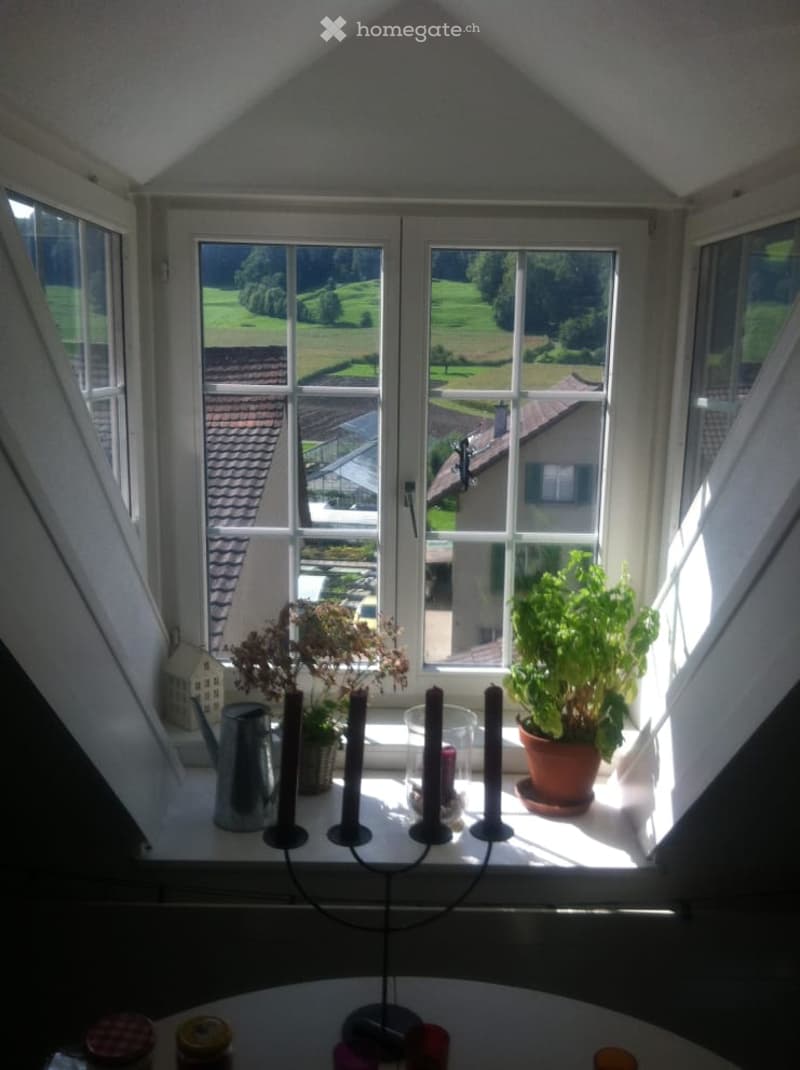 Bright attic room with large windows offering a scenic view of green fields and rooftops. The windowsill is decorated with a candle holder with multiple candles, a potted plant, and a teapot. A small table is visible in the foreground with various items o