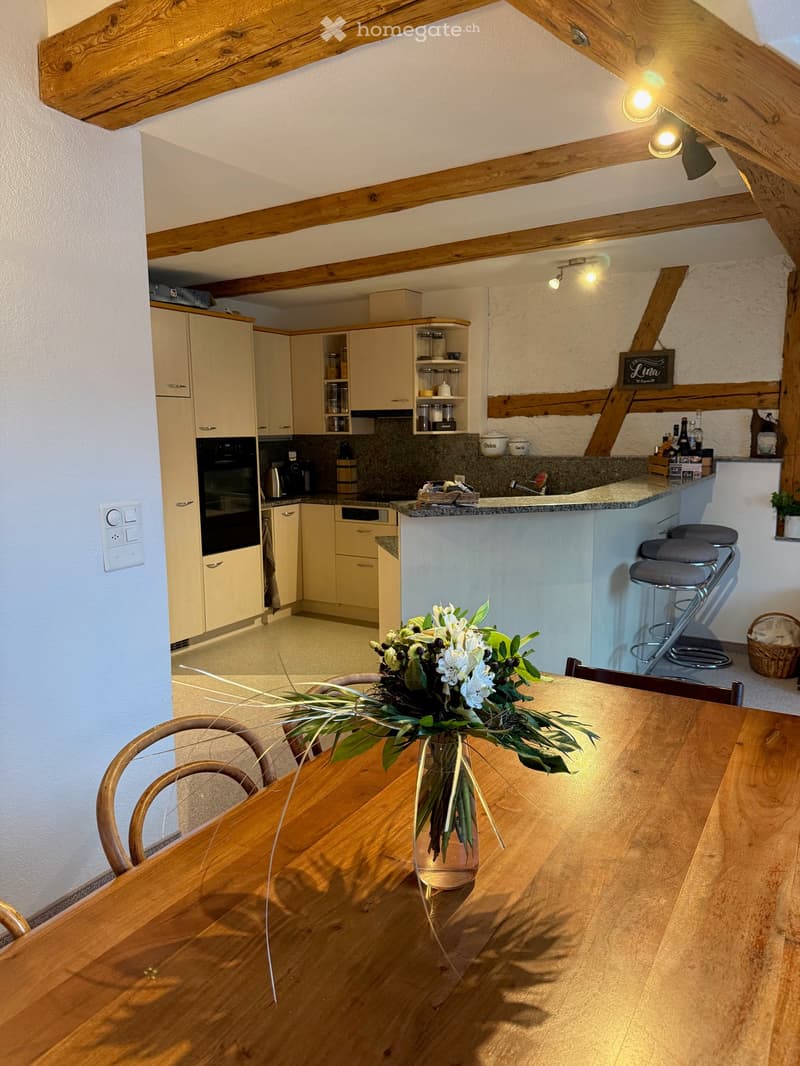 Bright kitchen with wooden beams, cream-colored cabinets, granite countertop, and a dining area with a wooden table and vase of flowers.