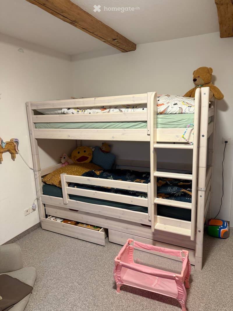 Cozy children's bedroom with a white wooden bunk bed, colorful bedding, stuffed animals, a pink playpen, and a toy on the floor.