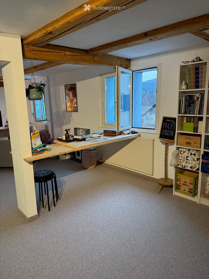 A cozy, well-lit room with exposed wooden beams, a large window, a wooden table with a laptop, a stool, a bookshelf filled with books, and a small plant hanging from the ceiling.