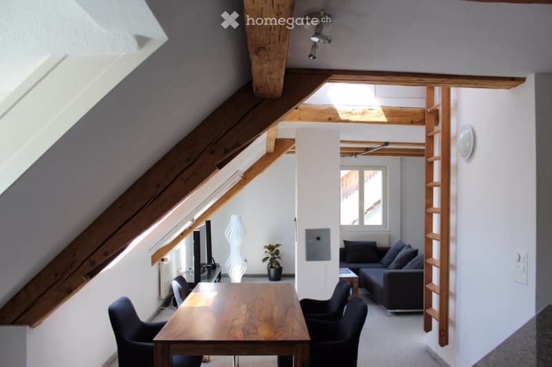 Bright attic living space with wooden beams, large windows, a sofa, dining table, chairs, a vase, and a potted plant.