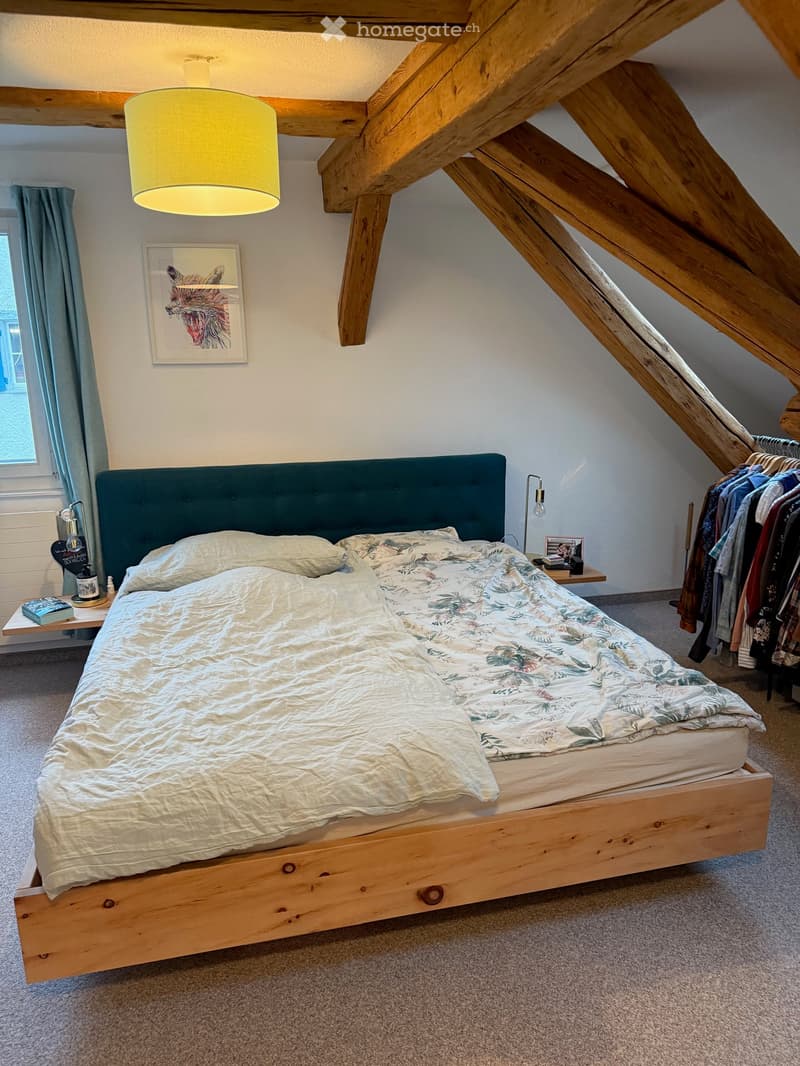 Cozy attic bedroom with wooden beams, a large bed with a teal headboard, a nightstand with a lamp, and a clothing rack with hanging clothes.