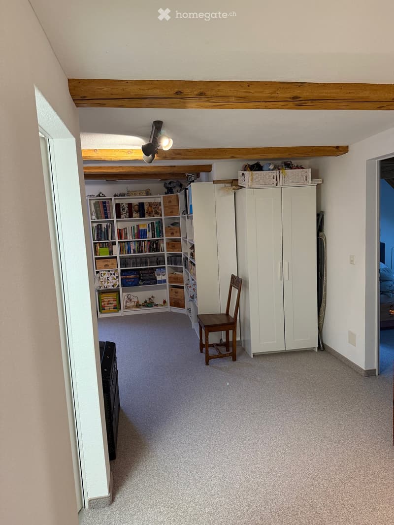 Carpeted room with exposed wooden beams, a wooden chair, a white cabinet, a bookshelf filled with books and boxes, and wicker baskets on top of the cabinet.