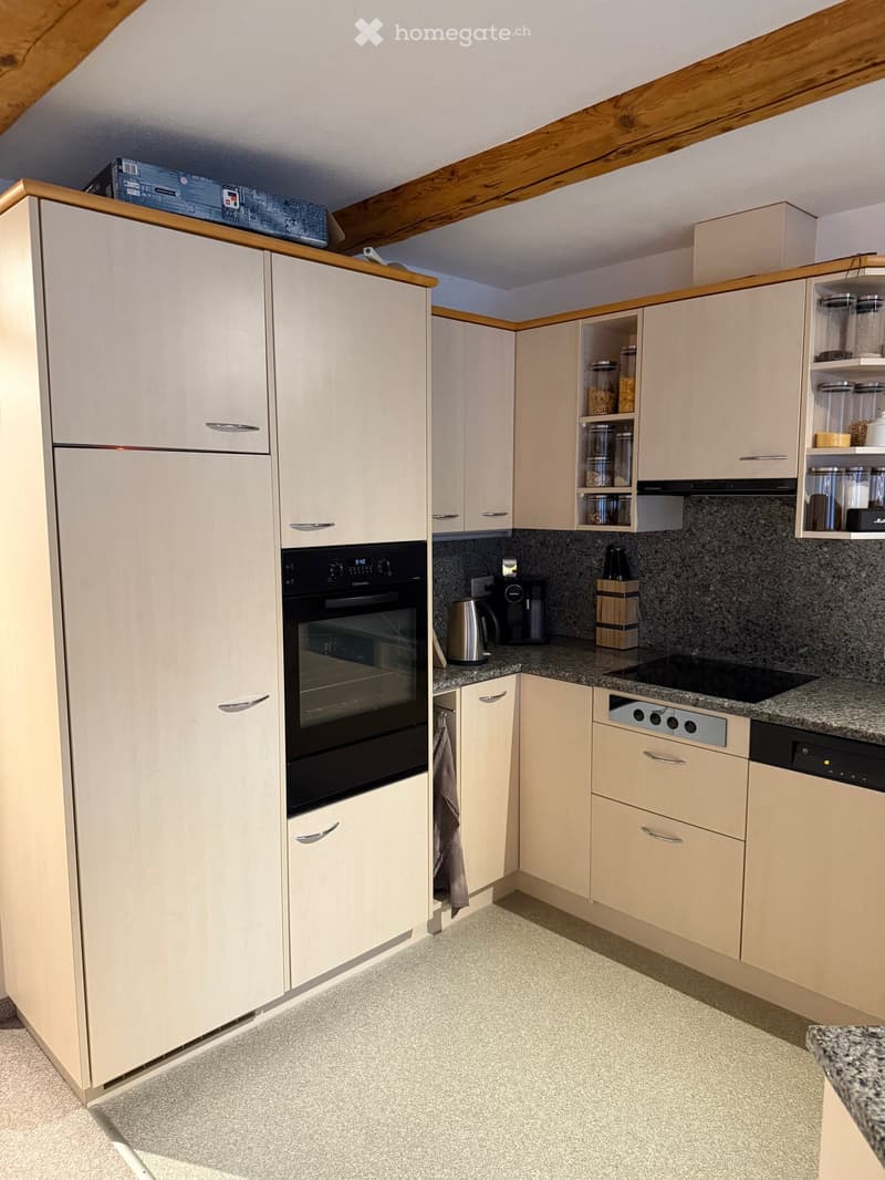 Modern kitchen with light-colored cabinets, stainless steel appliances, granite countertop, and a built-in oven. Storage jars and a coffee maker are visible on the counter.