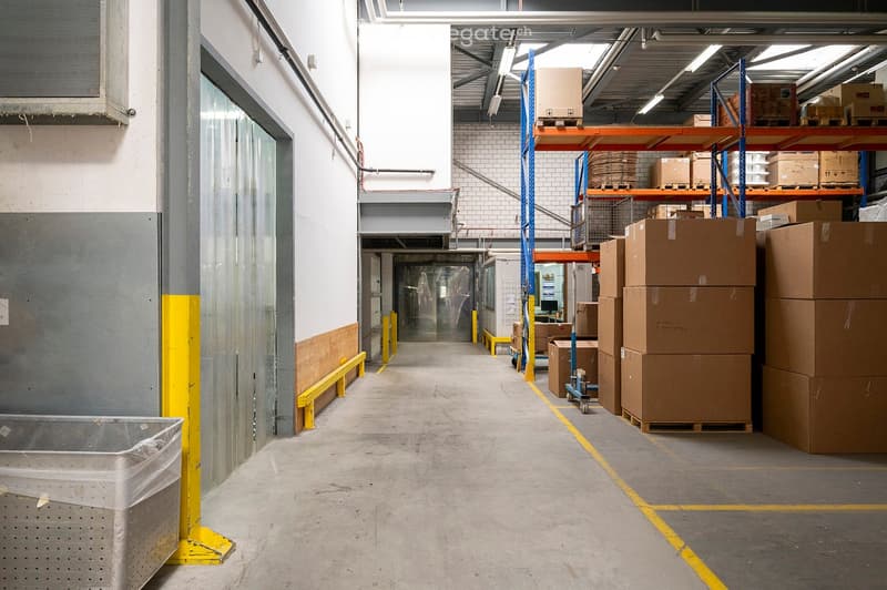 Warehouse interior with concrete floors, metal shelving units, stacked cardboard boxes, and industrial lighting.