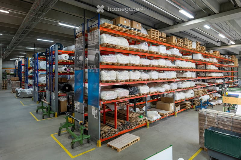 Warehouse with high ceilings, metal shelving units filled with various materials, including cardboard boxes, rolls of paper, and pallets. Machinery and equipment are visible, with a clean and organized layout.