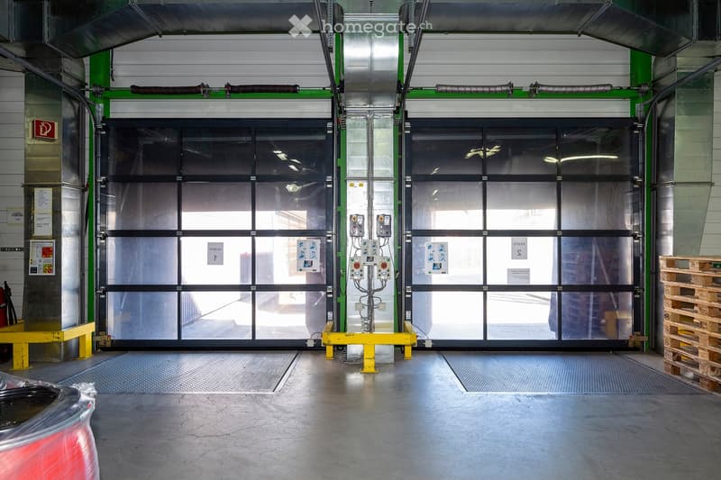 Empty industrial warehouse space with large roll-up doors, metal floor, and control panels visible.