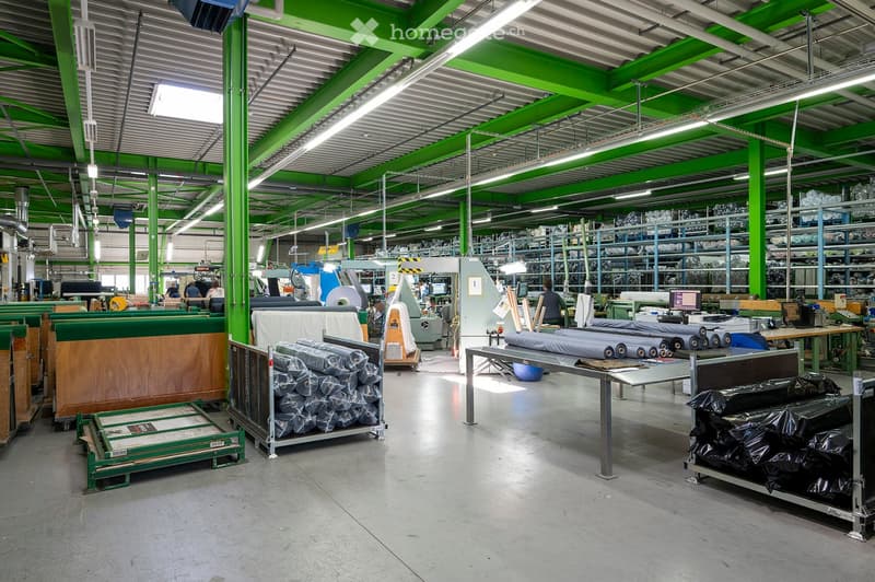 Large industrial warehouse with green structural beams, metal shelving units, and various machinery. The space features a high ceiling with exposed lighting and ventilation ducts, with rolls of material and equipment visible throughout.