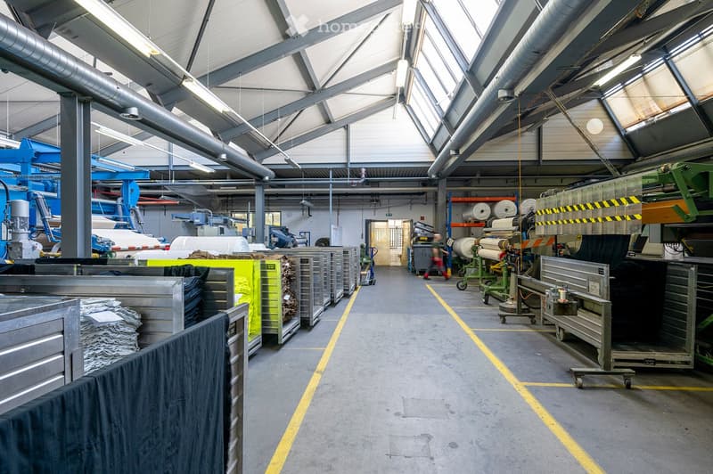 Large industrial warehouse with high ceilings, metal beams, and various machinery and equipment, including large rolls of material and storage racks.