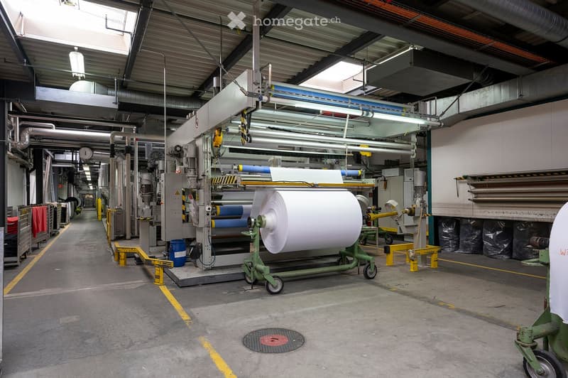 Large industrial factory interior with machinery, conveyor belts, and a large roll of paper on a cart. The space features a high ceiling with exposed metal beams, industrial lighting, and various equipment lining the walls.