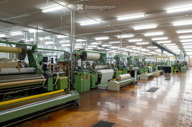 Industrial factory space with large green textile machines, rolls of fabric, and a polished wooden floor.