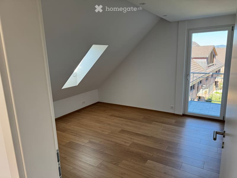 Empty attic room with wooden flooring, white walls, skylight window, and sliding glass door leading to a balcony.