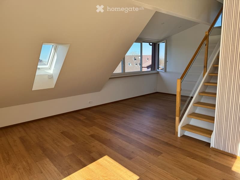 Empty attic room with wooden flooring, slanted ceilings, and a staircase leading upwards. Features large windows and skylights allowing natural light to enter.