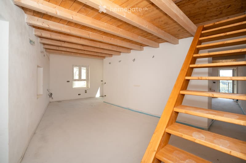 Empty room with exposed wooden beams on the ceiling, a wooden staircase leading upwards, and a small window letting in natural light. The walls are white, and the floor is unfinished concrete.