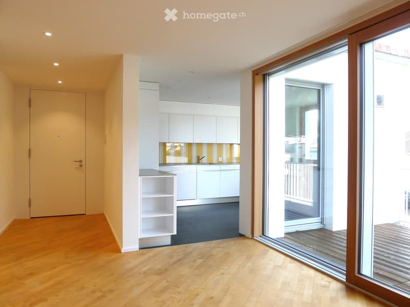 Modern, minimalist kitchen with light grey cabinets, a sink, and a small shelf. The room features wooden flooring and large glass doors leading to a balcony.